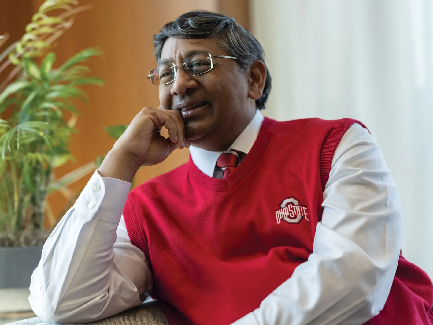 Ravi Bellamkonda leans one elbow on the arm of a couch as he leans into it and chuckles. He wears a scarlet Ohio State sweater vest, glasses and has salt-and-pepper tussled hair. He appears to be invested in a conversation