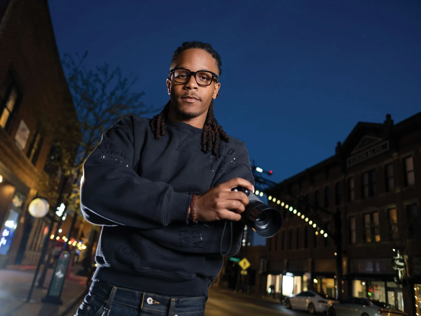 Justen Turner is a young black man wearing thick-framed glasses and a sweatshirt. He looks focused as he poses for this portrait holding a small video camera. His hair is dreads pulled back in a low ponytail. He looks like he's competent, professional and thoughtful—or like someone you'd trust to make a movie about yourself or a loved one. He's photographed in the Short North after the sun has set.