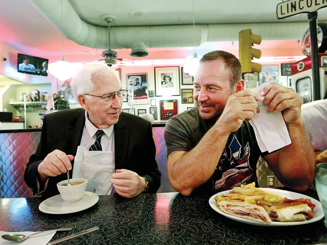 This image shows two people seated next to each other at a diner-style booth, engaged in conversation while eating. Hugh Dorrian is an older white man with white hair and a friendly, grandfatherly smile. He has a small bowl of soup. The other man is younger and wears a T-shirt and has a sandwich and fries.