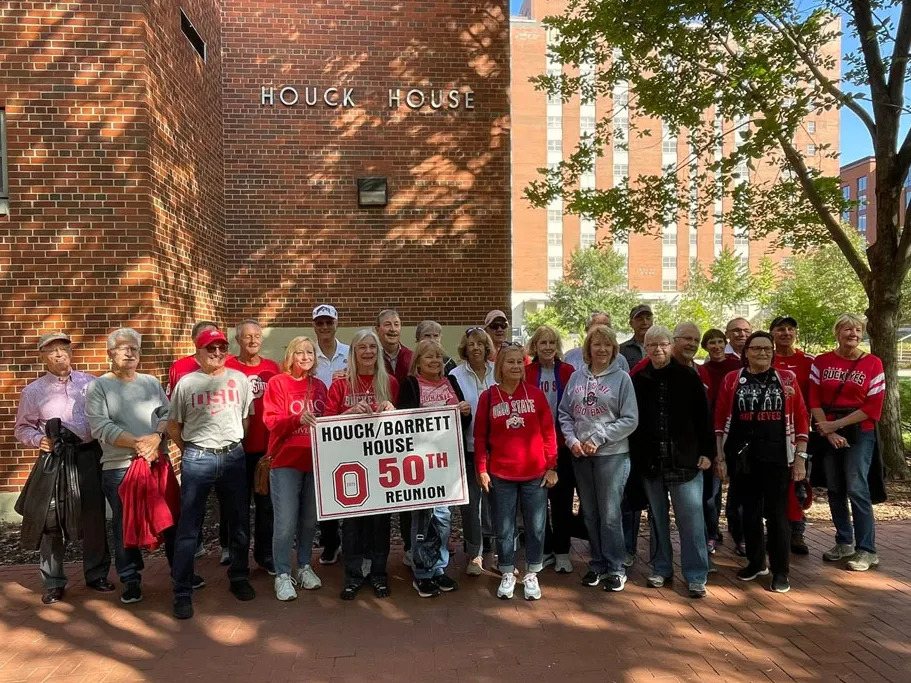 About 25 people pose for a photo with a big sign that says Houck/Barrett House 50th reunion. They stand in front of the dormitory Houck House, which is red brick. The people wear Ohio State gear, and the sun shines.