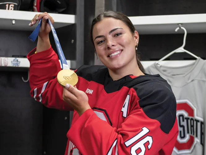 Joy Dunne smiles as she proudly shows off her gold medal in the locker room for Ohio State women's hockey. She wears her team uniform. 
