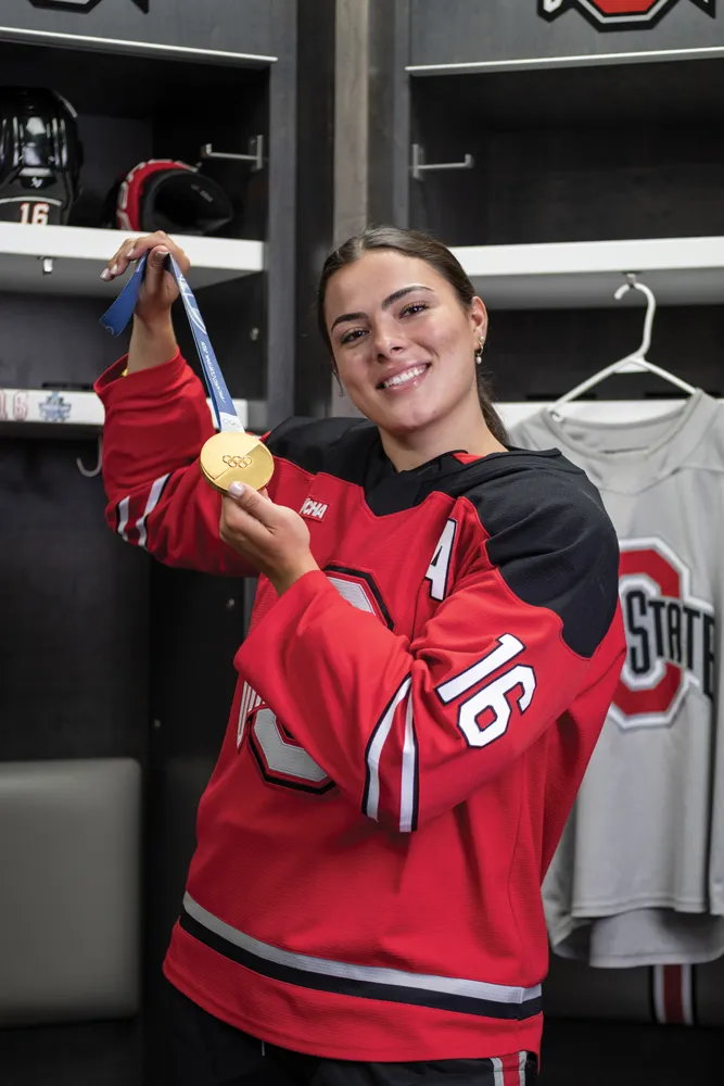 Joy Dunne smiles as she proudly shows off her gold medal in the locker room for Ohio State women's hockey. She wears her team uniform. 