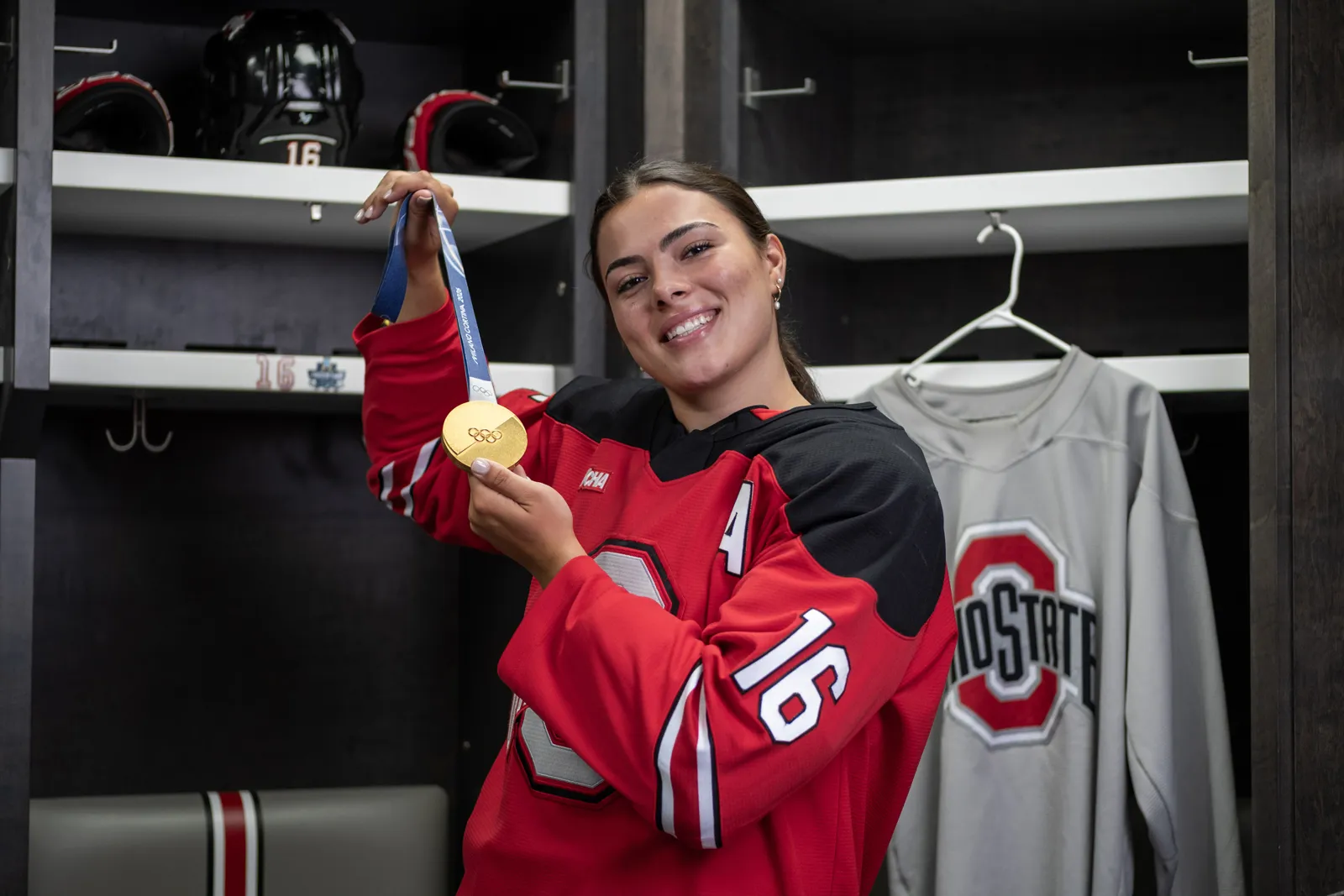 Joy Dunne smiles as she proudly shows off her gold medal in the locker room for Ohio State women's hockey. She wears her team uniform. 