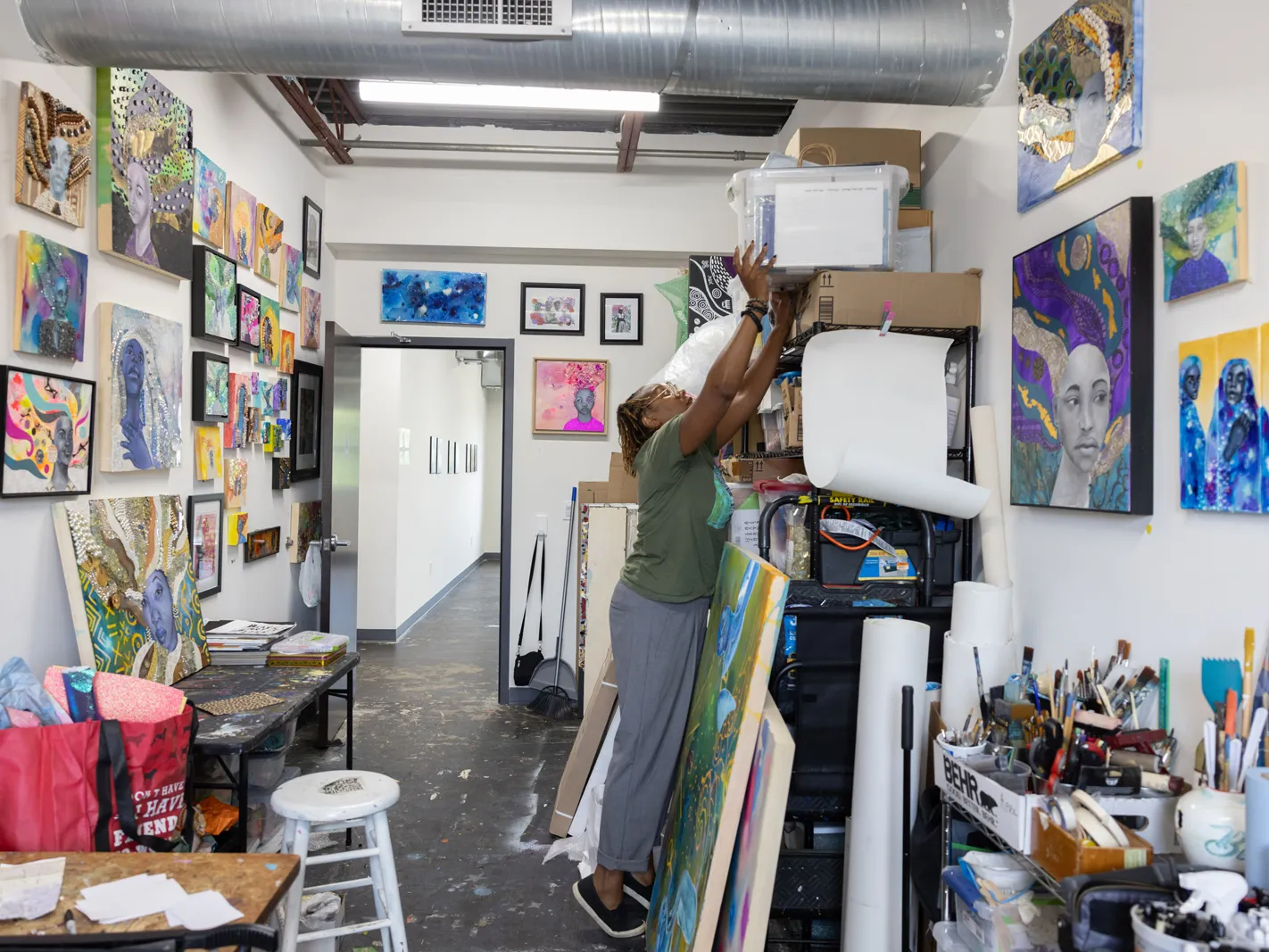 Sunami reaches higher than she is tall for a plastic bin on top of shelves and other stacks of supplies. She's in her studio which is full of interesting paintings and tools 