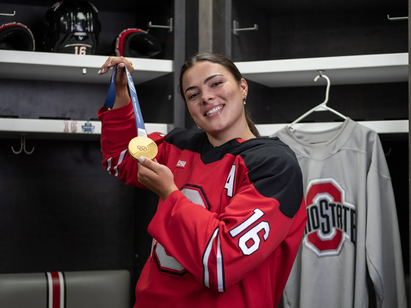Joy Dunne smiles as she proudly shows off her gold medal in the locker room for Ohio State women's hockey. She wears her team uniform. 
