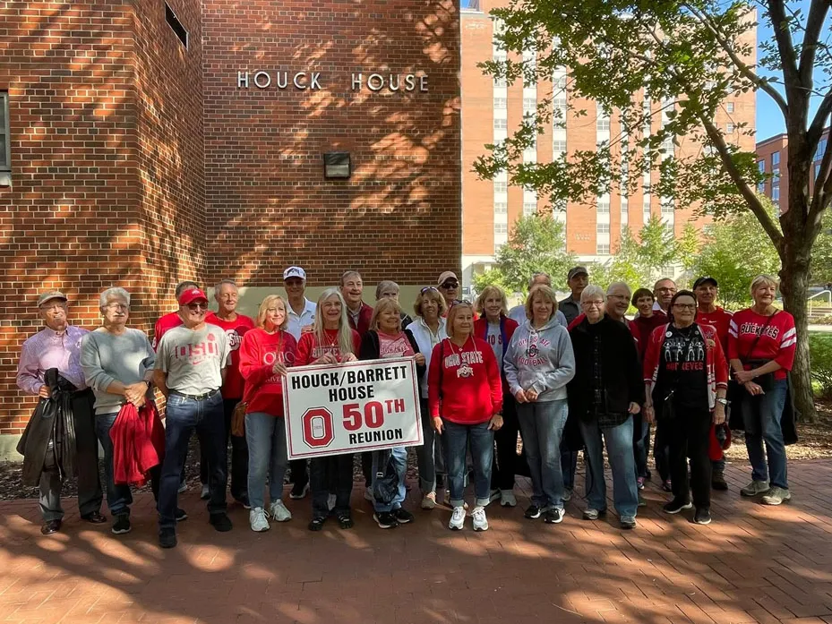 About 25 people pose for a photo with a big sign that says Houck/Barrett House 50th reunion. They stand in front of the dormitory Houck House, which is red brick. The people wear Ohio State gear, and the sun shines.