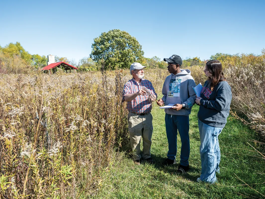 Holding a plant, a professor talks to two students taking notes on the prairie at Ohio State Marion.