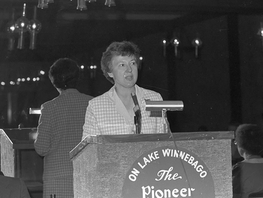 Helen Briwa speaks at a podium in an old black and white photo. She wears a checked jacket and a mirror behind her reflects her back and, faintly, the audience.