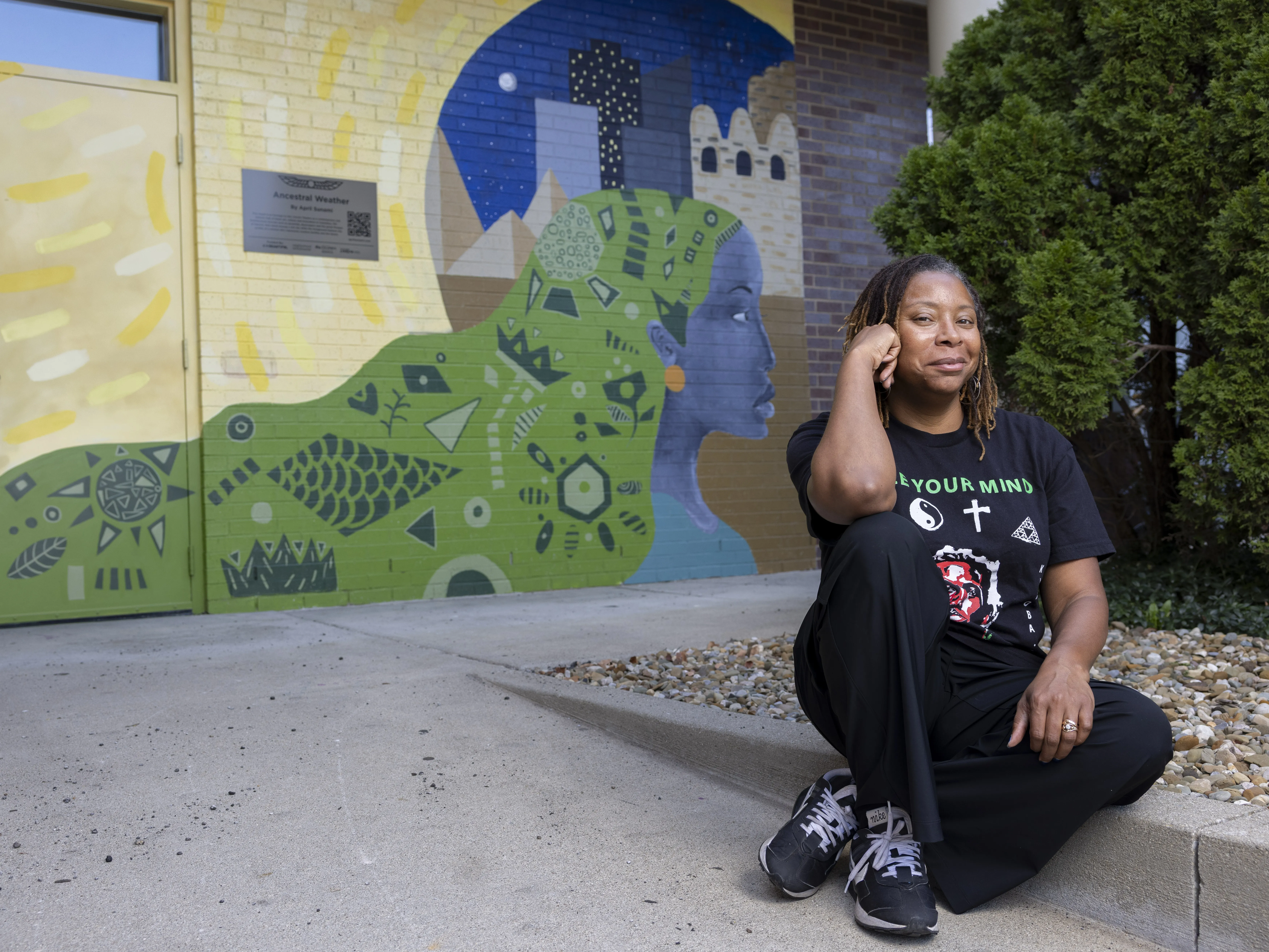 April Sunami smiles knowingly as she sits on a curb in front of a colorful mural she painted