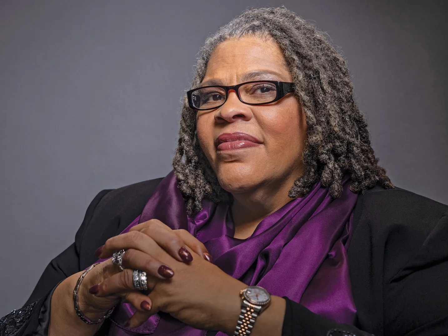 Patrice Palmer seems proud, thoughtful and friendly but direct as she poses for this photo with a slight smile that nevertheless lights up her face. She seems to be looking the viewer in the eye. She is an older Black woman with shoulder-length hair and wears rectangular glasses, a business suit, a silk scarf and silver rings and watch. 