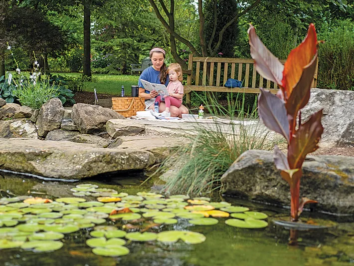 A young mother reads to her child alongside a pond with lilly pads and interesting plants