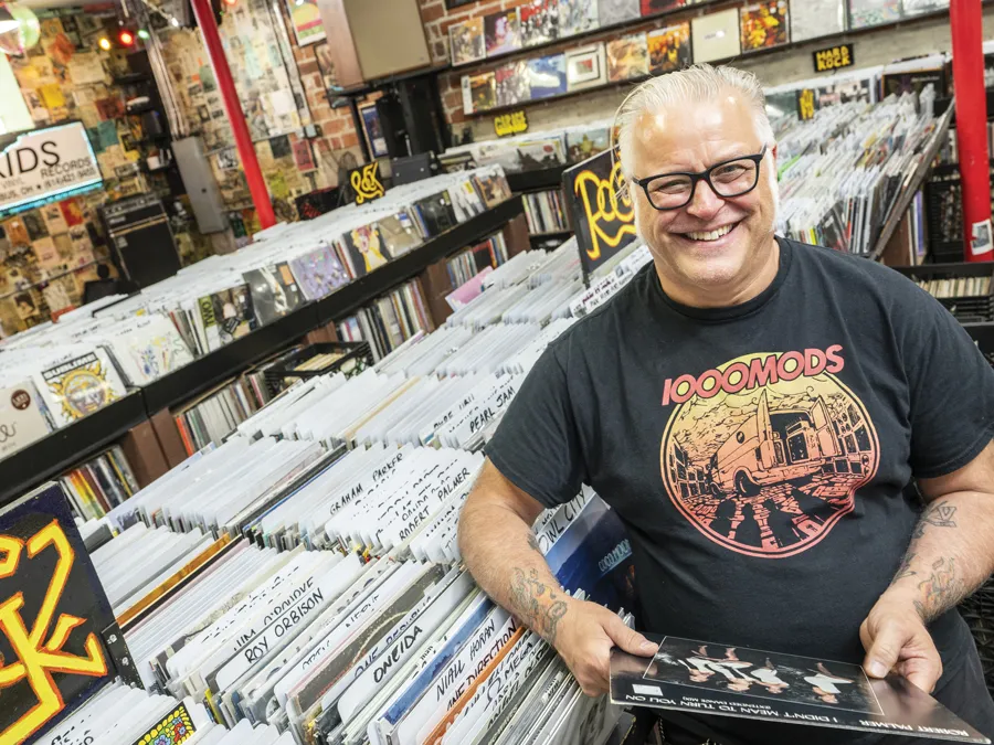 Used Kids owner Greg Hall laughs as he is photographed in the record store, wearing thick-framed glasses and an artful T-shirt for the hard rock band 1000 Mods.