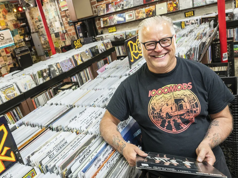 Used Kids owner Greg Hall laughs as he is photographed in the record store, wearing thick-framed glasses and an artful T-shirt for the hard rock band 1000 Mods.