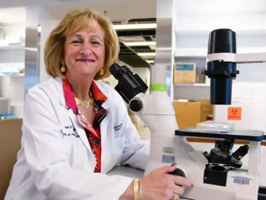 Gail Besner smiles as she looks up from her microscope while wearing a lab coat.