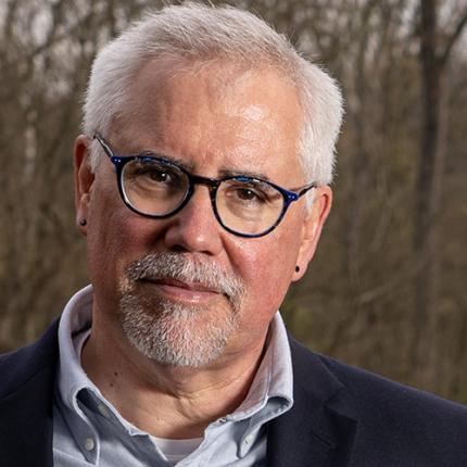 A man whose soft smile is reflected in his lips and eyes poses for a photo in the woods. He wears round glasses and a button-up under a suit coat, whose color is subtly reflected in the frames of his glasses