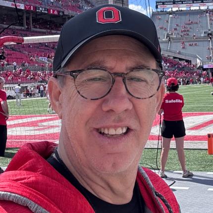 Mike Larsen smiles while on the sideline of Ohio Stadium. He wears an Ohio State hat and sweatshirt, as well as round glasses.