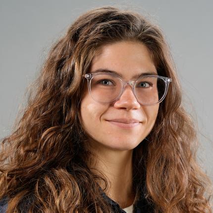 Mattie Mueller softly smiles as poses for a portrait. She has long curly hair and clear-rimmed glasses